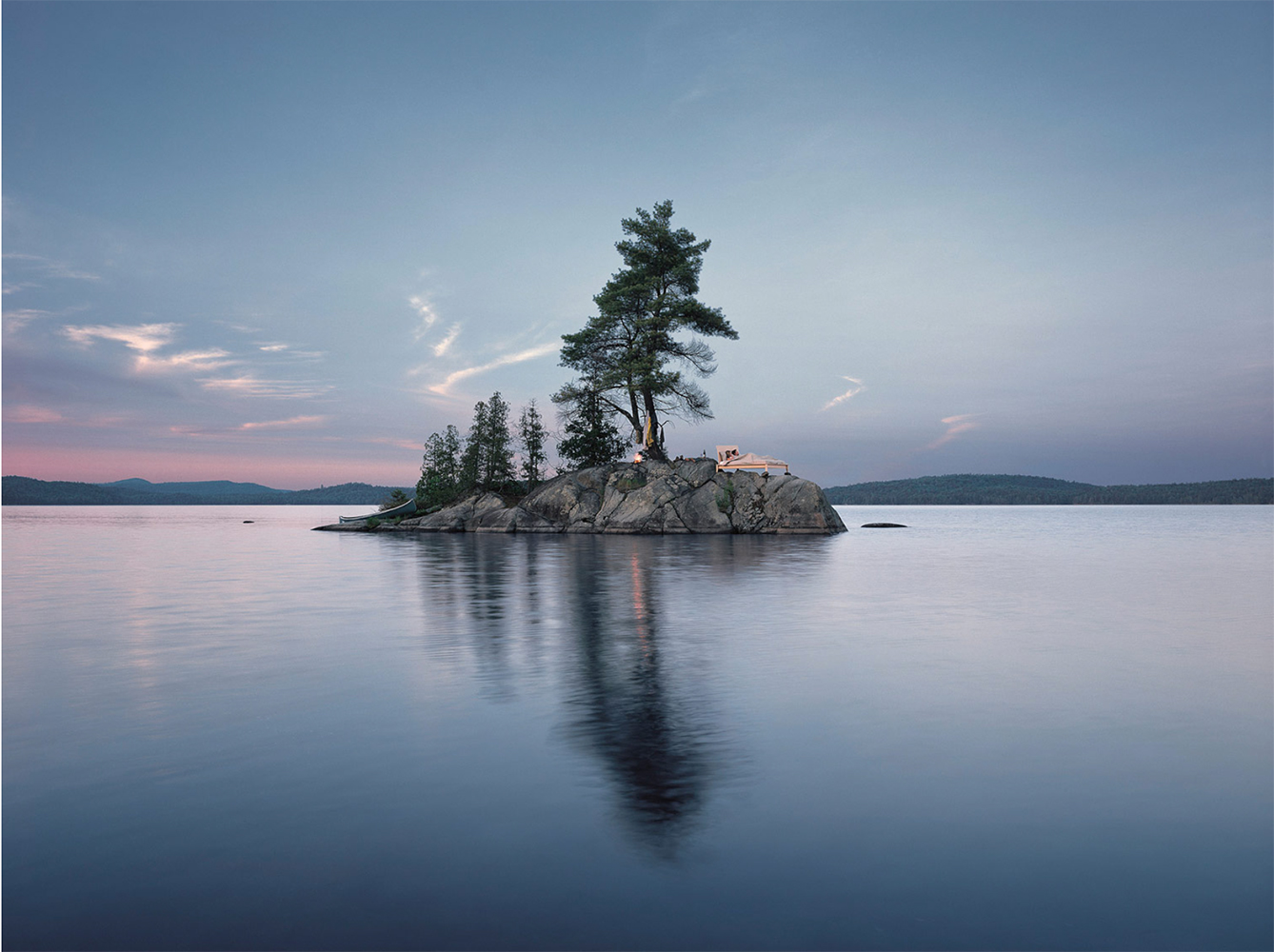 Still lake reflects a rocky island with trees at dusk. A small structure sits on the island illuminated by warm interior light.