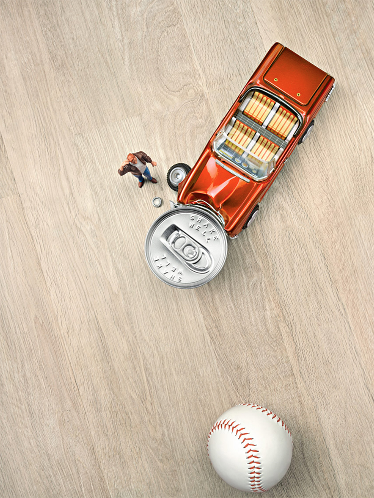 Miniature car crash scene featuring a toy car atop a crushed soda can with a tiny figure nearby, set on a light wooden surface.