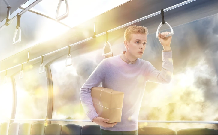 Young adult male stands on a bus with a package, holding onto a handrail, wearing a light sweater. Bright, sunlit scene.