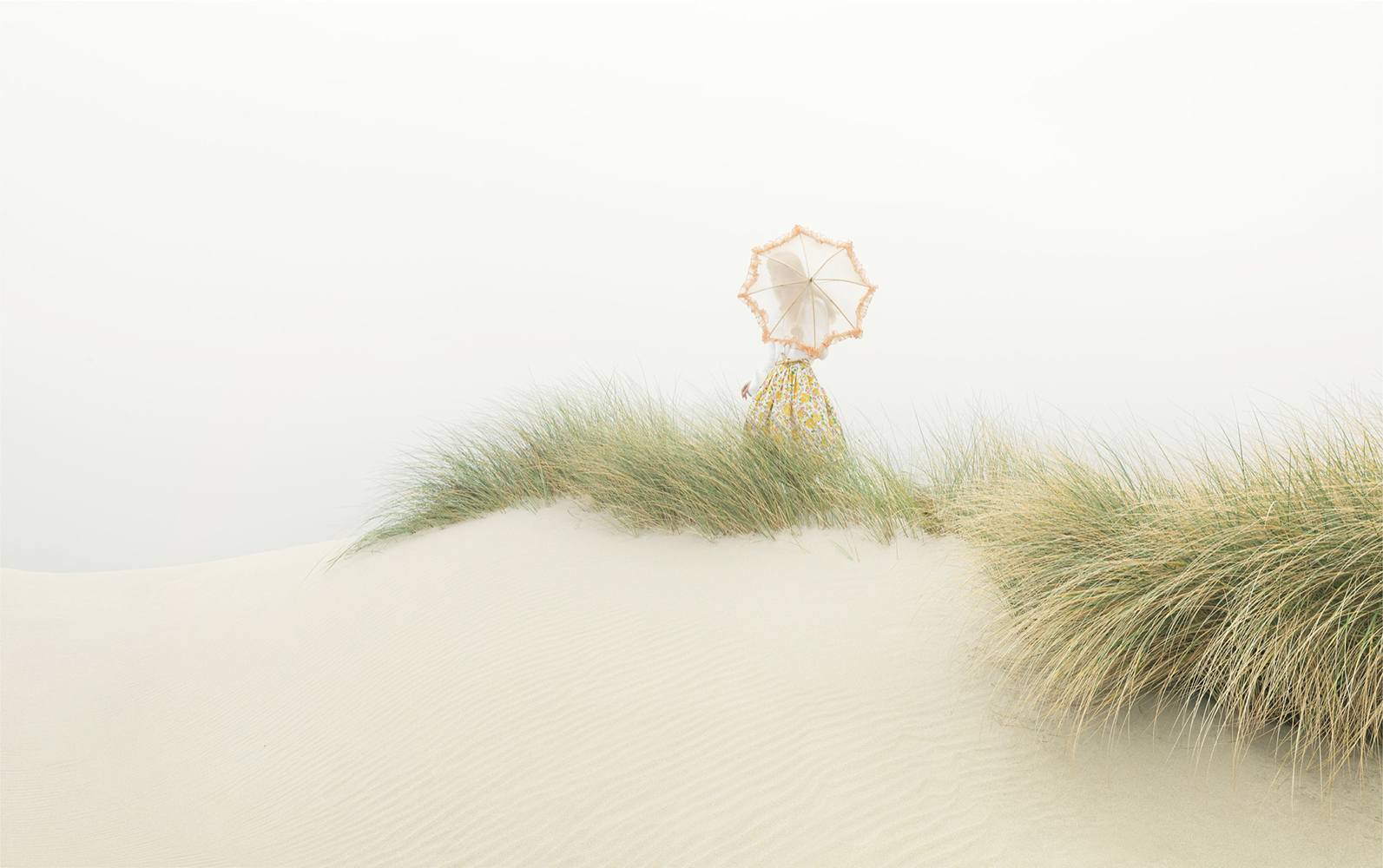 Woman in a patterned skirt stands on a sand dune holding an umbrella, surrounded by beach grass in a muted, hazy outdoor setting.