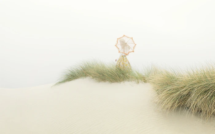 Figure holding a patterned parasol standing among sandy dunes and sea grass under an overcast sky.