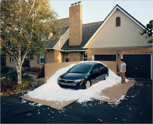 A black sedan surrounded by packing peanuts on cardboard panels in a suburban driveway in front of a house.