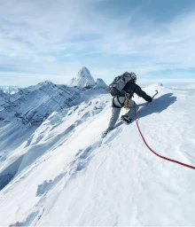 Climber wearing a harness and backpack ascending a steep snowy mountain ridge with an ice axe in hand.