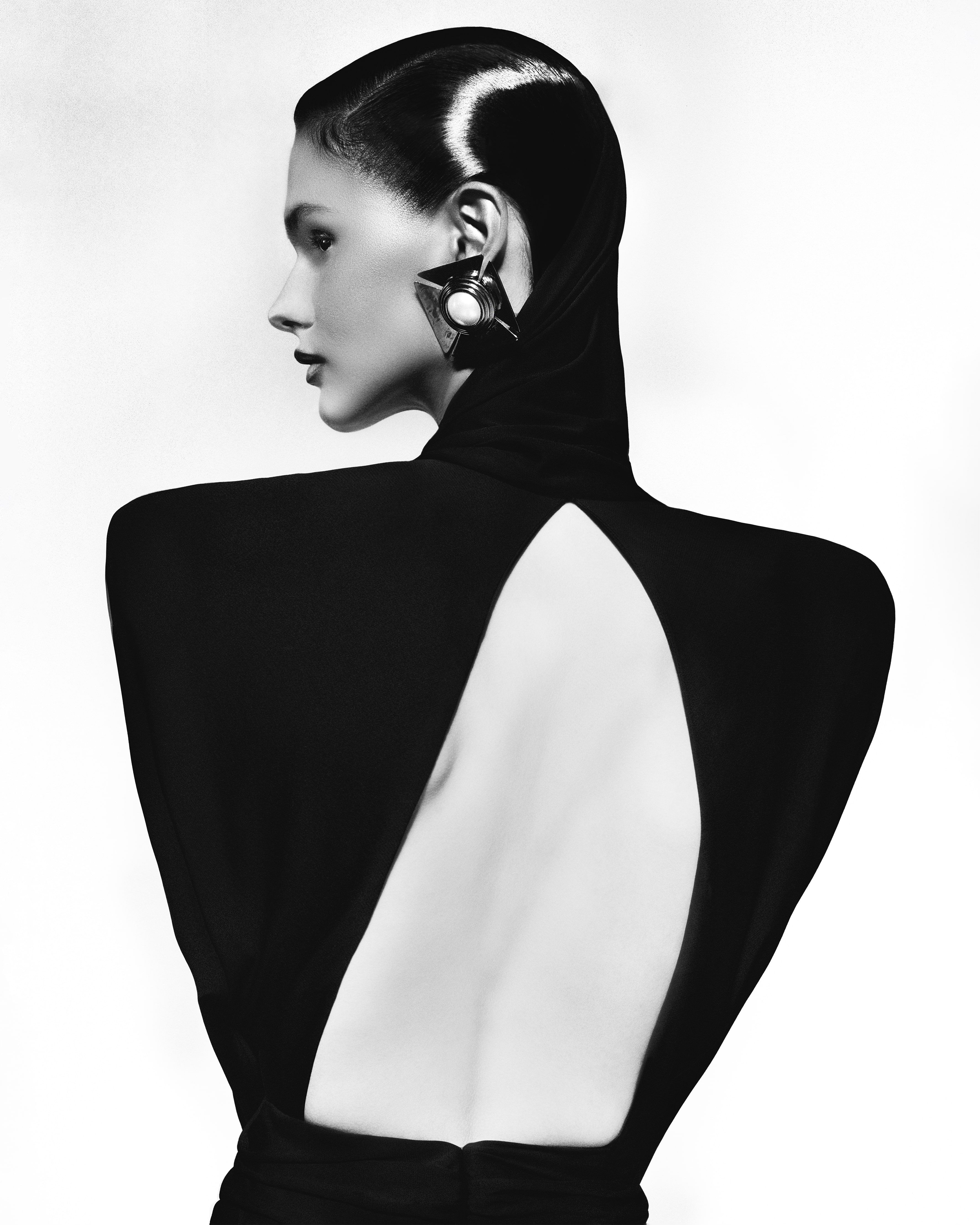Elegant black-and-white portrait of a woman with a slicked-back hairstyle, prominent earring, and a black turtleneck dress with open back.