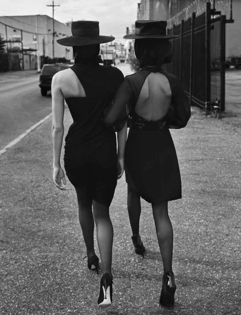 Two women walk arm in arm down a city street in black dresses, stockings, and heels, with matching wide-brimmed hats, from a back view.