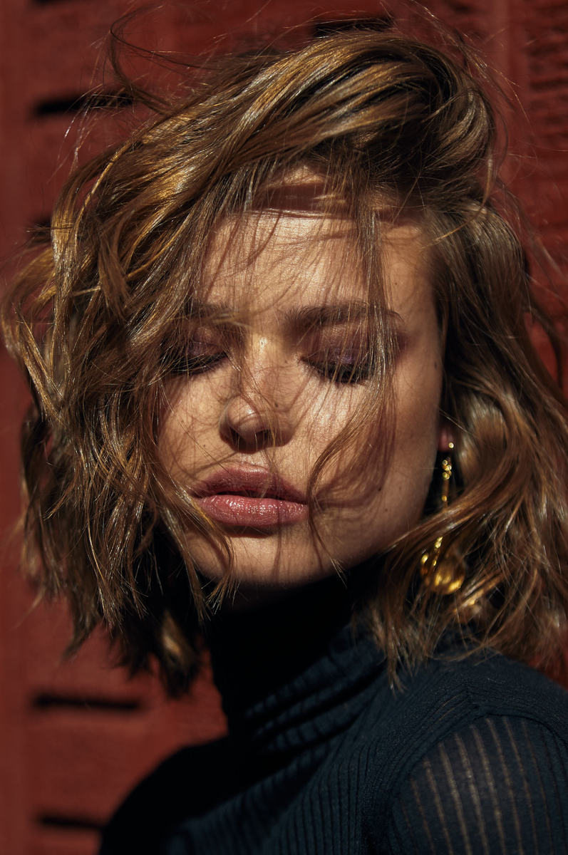 Close-up portrait of a young adult woman with short, tousled brown hair and golden earrings, wearing a black turtleneck sweater.