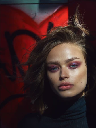 Portrait of a beautiful young woman with professional purple eye makeup, soft pink lipstick, and naturally tousled hair, against a red background.