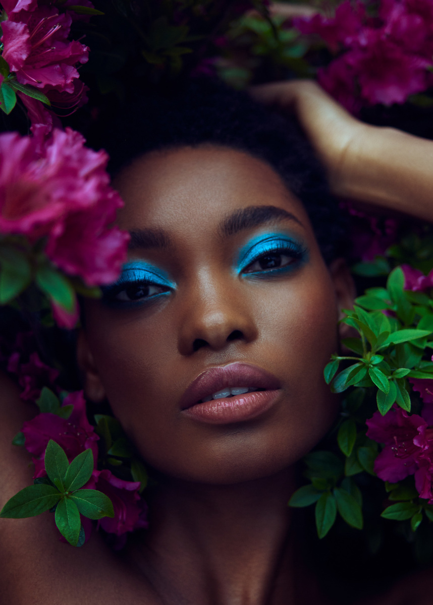 Portrait of a Black woman with vibrant blue eyeshadow surrounded by deep pink flowers