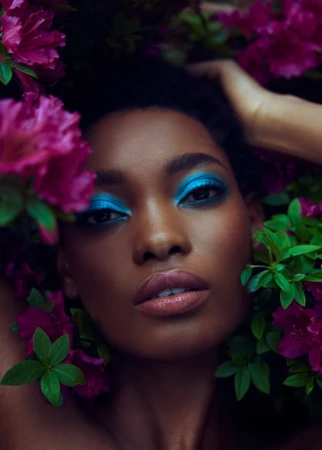 Portrait of a Black woman with vibrant blue eyeshadow surrounded by deep pink flowers