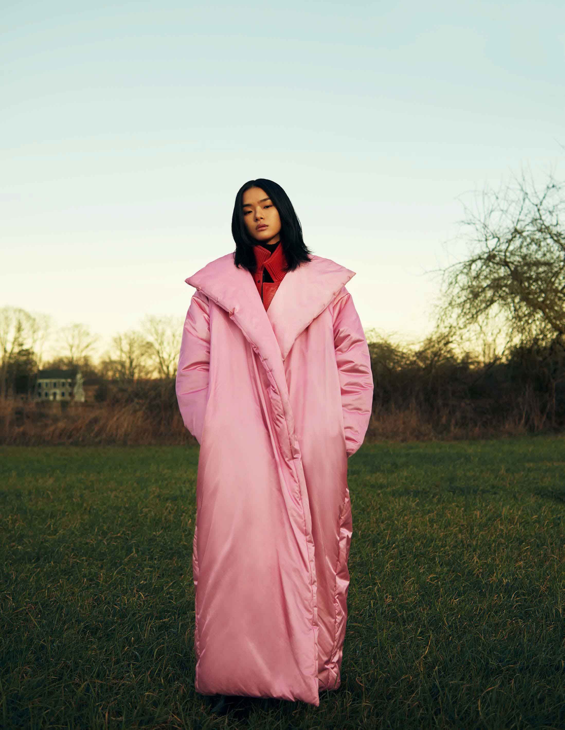A stylish woman in a pink puffer coat stands in a grassy field beneath a pale sky, blending fashion and nature with elegance and warmth.
