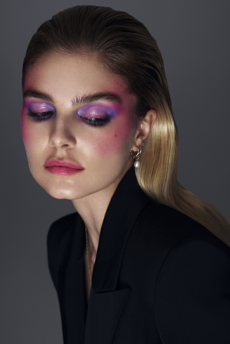 Portrait of a fair-skinned woman with wet blonde hair and striking purple and pink makeup, wearing a black blazer, on a gray background.
