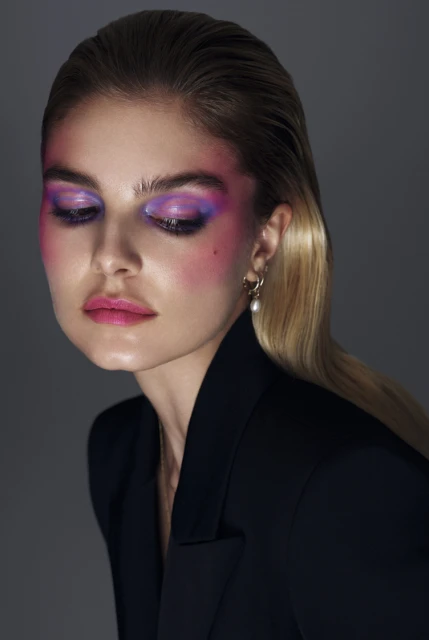 Portrait of a fair-skinned woman with wet blonde hair and striking purple and pink makeup, wearing a black blazer, on a gray background.
