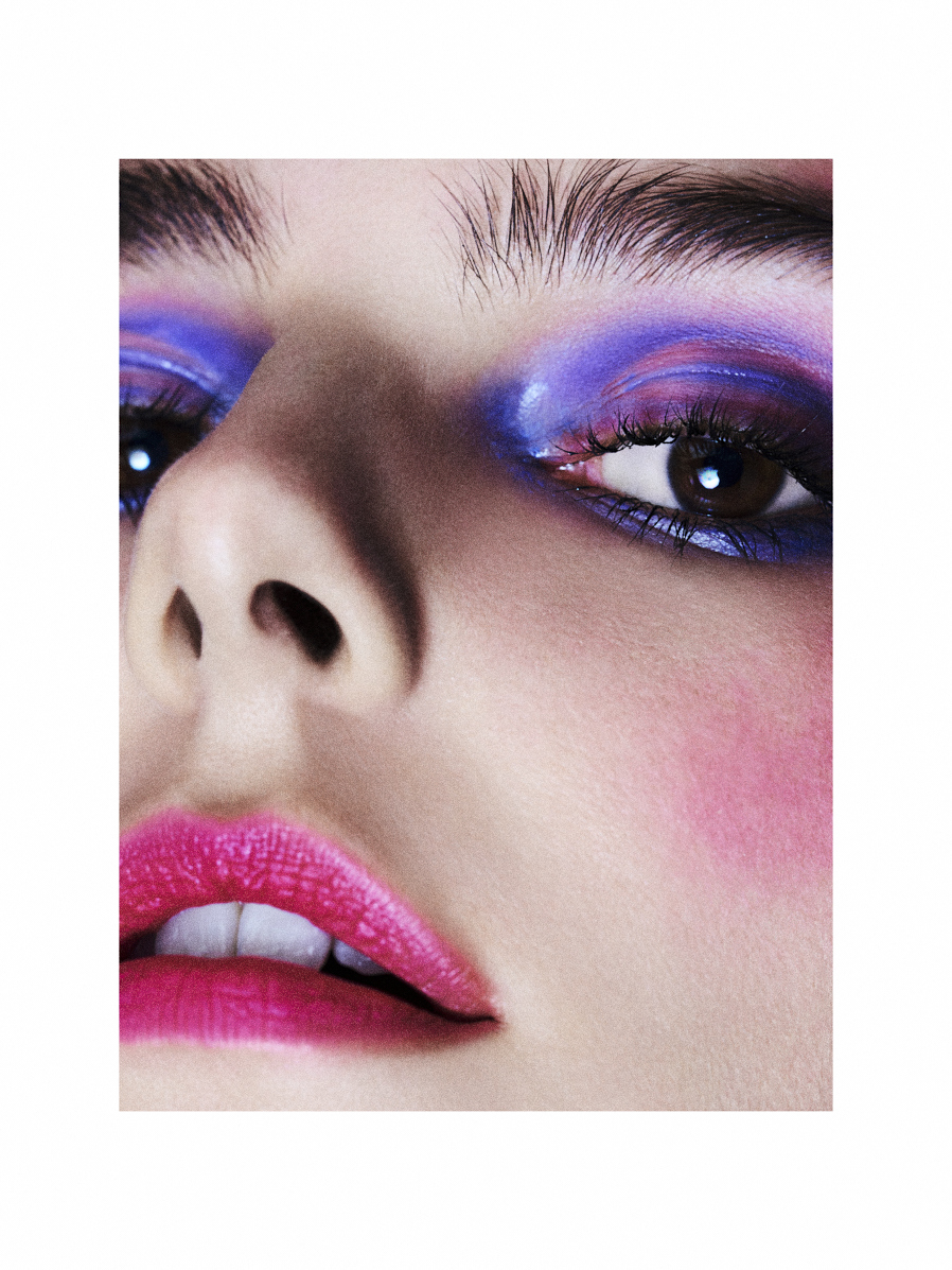 Close-up of a woman's face with vibrant makeup featuring blue and purple eyeshadow and bold pink lipstick, set against a neutral background.