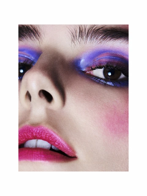 Close-up of a woman's face with vibrant makeup featuring blue and purple eyeshadow and bold pink lipstick, set against a neutral background.
