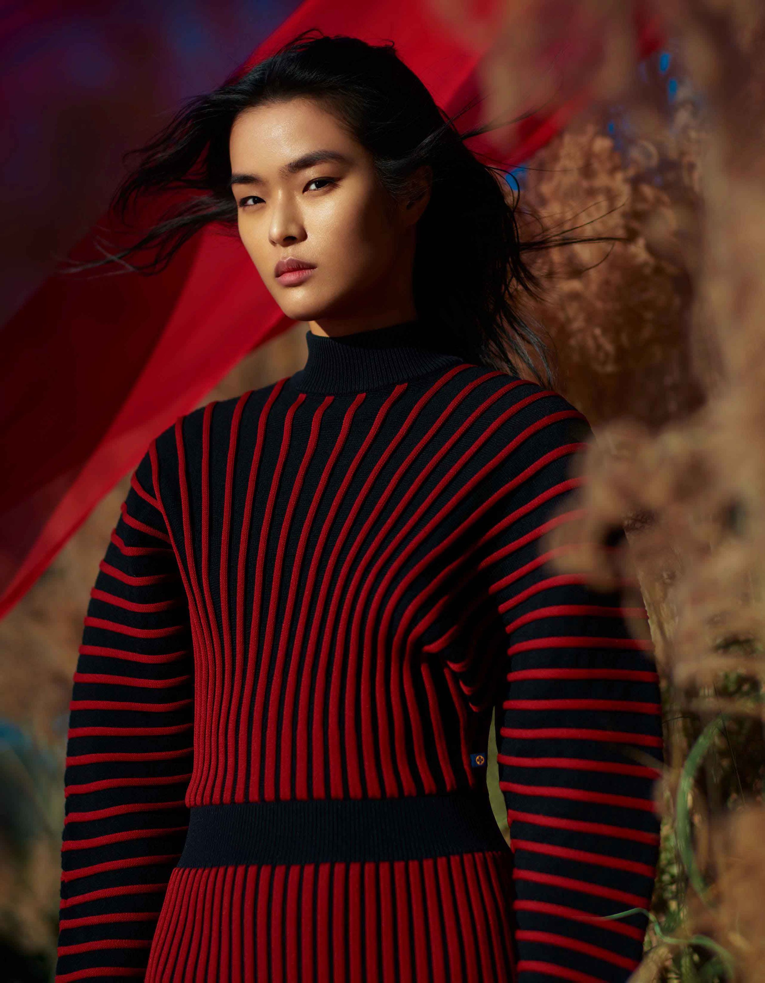 Asian woman with dark hair poses in a red and black striped sweater and dress, set against a backdrop of grass and red fabric.