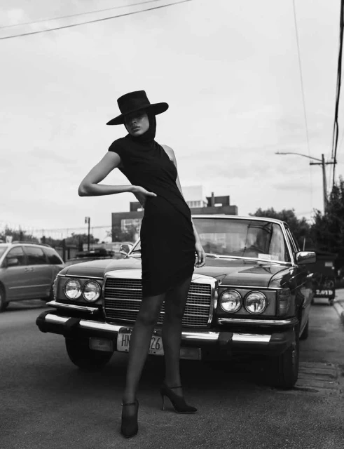 Monochrome fashion portrait of a woman in a black dress and wide-brimmed hat posing near a classic car on a city street.