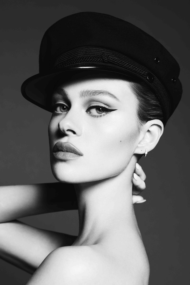 Monochrome portrait of a young woman with bold makeup, wearing a black hat, posing in a studio with a grey backdrop and soft lighting.