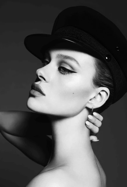 Black and white portrait of a woman wearing a black hat and dark eye makeup, posing with hand near neck, studio lighting, grey background.