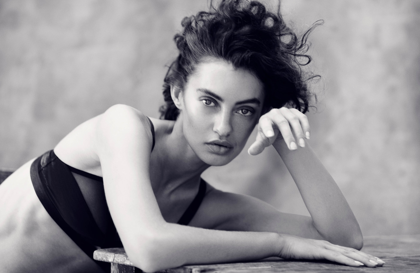 Monochrome portrait of a woman with dark curly hair, gazing directly at the camera while resting her arms on a wooden surface.