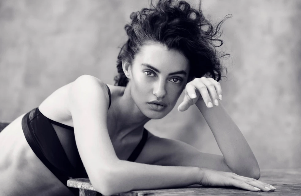 Monochrome portrait of a woman with dark curly hair, gazing directly at the camera while resting her arms on a wooden surface.