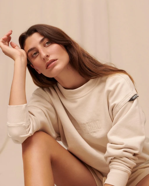 Portrait of a young woman in a beige sweatshirt. She rests her head on her hand, and looks directly at the camera with a neutral expression.