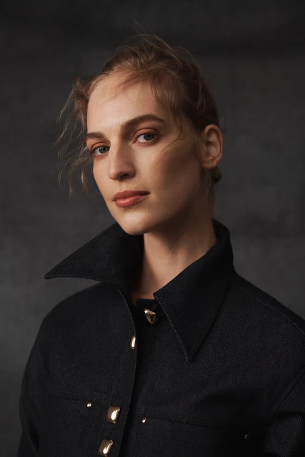 Close-up studio portrait of a fair-skinned adult woman wearing a dark collared denim jacket with gold buttons against a gray backdrop.