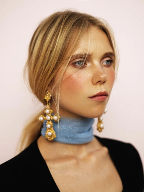 Close-up portrait of a blonde woman with blue eyes wearing statement earrings and a pearl-encrusted choker against a light pink background.