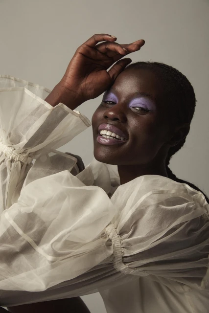 Portrait of a smiling Black woman with lavender eyeshadow and glossy lipstick, wearing a semi-sheer ruffled white blouse against grey.