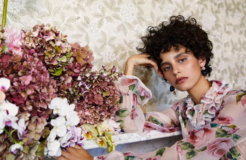 Close-up fashion portrait of a curly-haired woman in a floral dress, resting her head near a pink hydrangea flower arrangement.