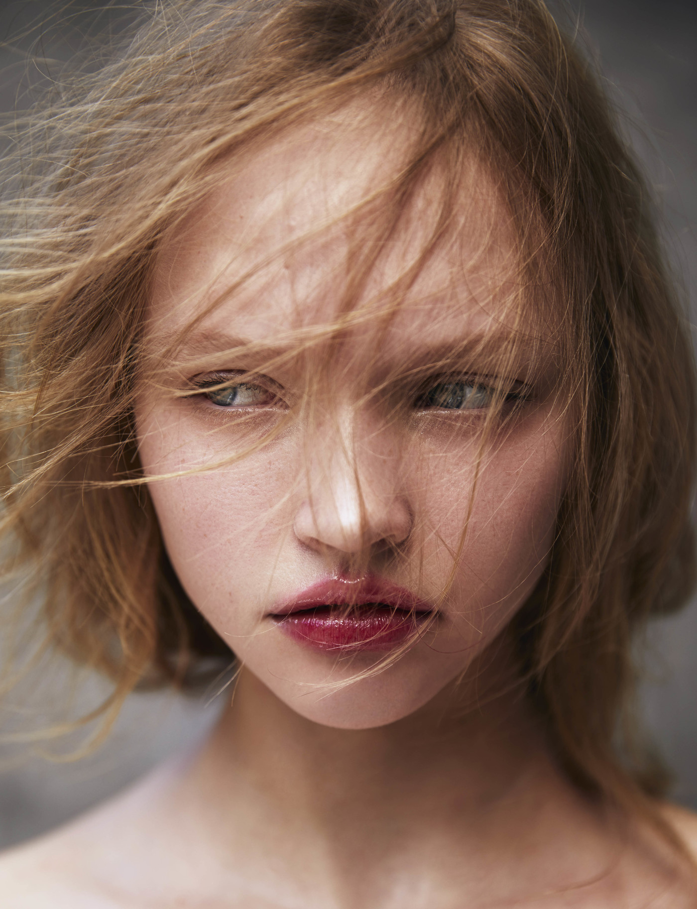 Close-up portrait of a woman with blonde hair and red lipstick, gazing off to the side with a blurred neutral background. Fashion photography.
