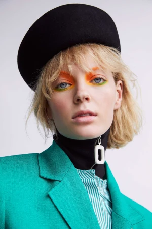 Model wearing a black beret, teal blazer, and vibrant orange and green eye makeup against a white backdrop.