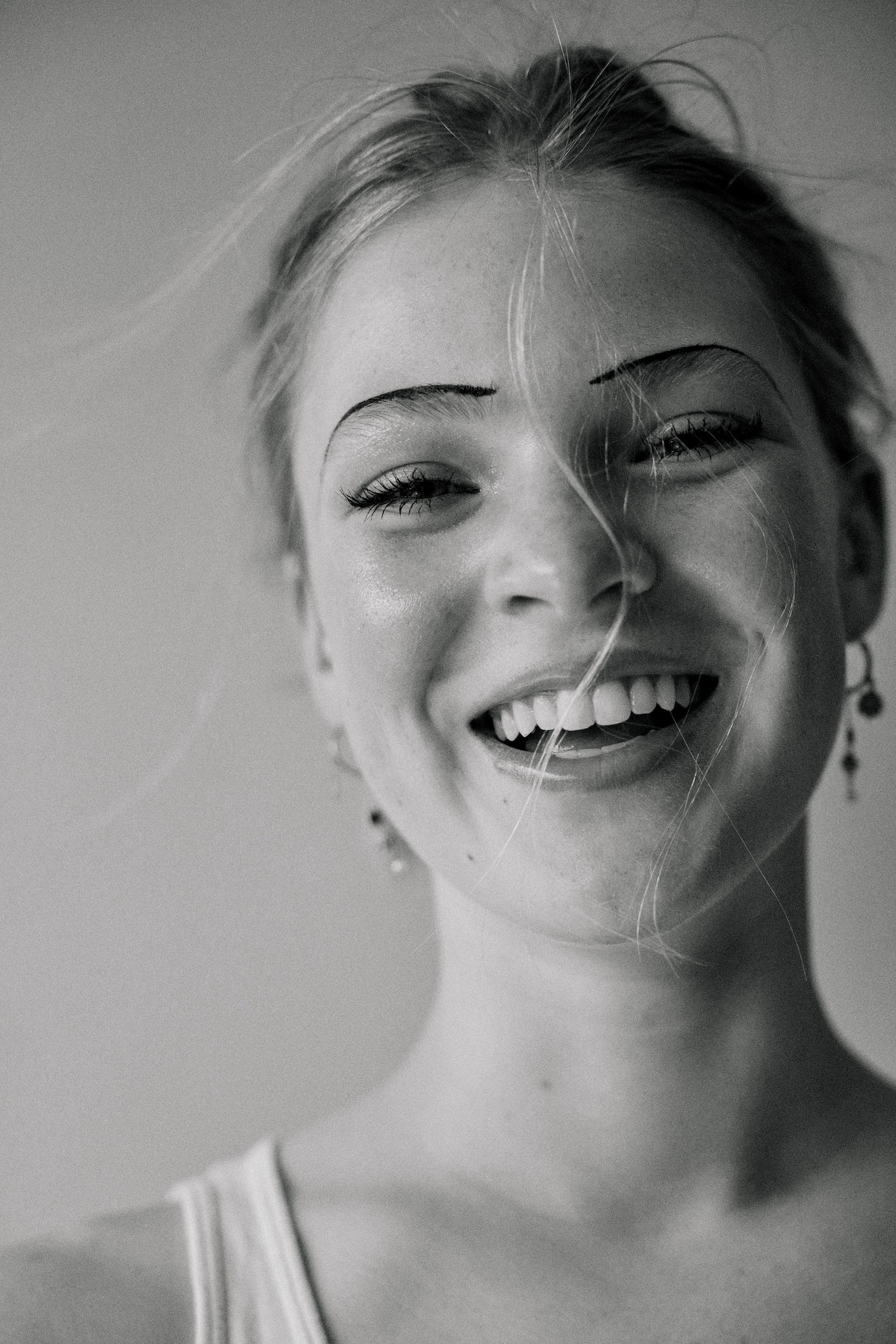 Close-up monochrome portrait of a smiling young woman with thin arched eyebrows and loose hair strands across her face