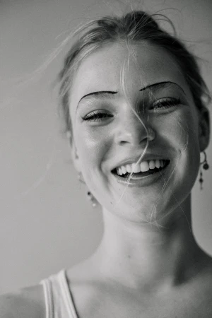 Close-up monochrome portrait of a smiling young woman with thin arched eyebrows and loose hair strands across her face