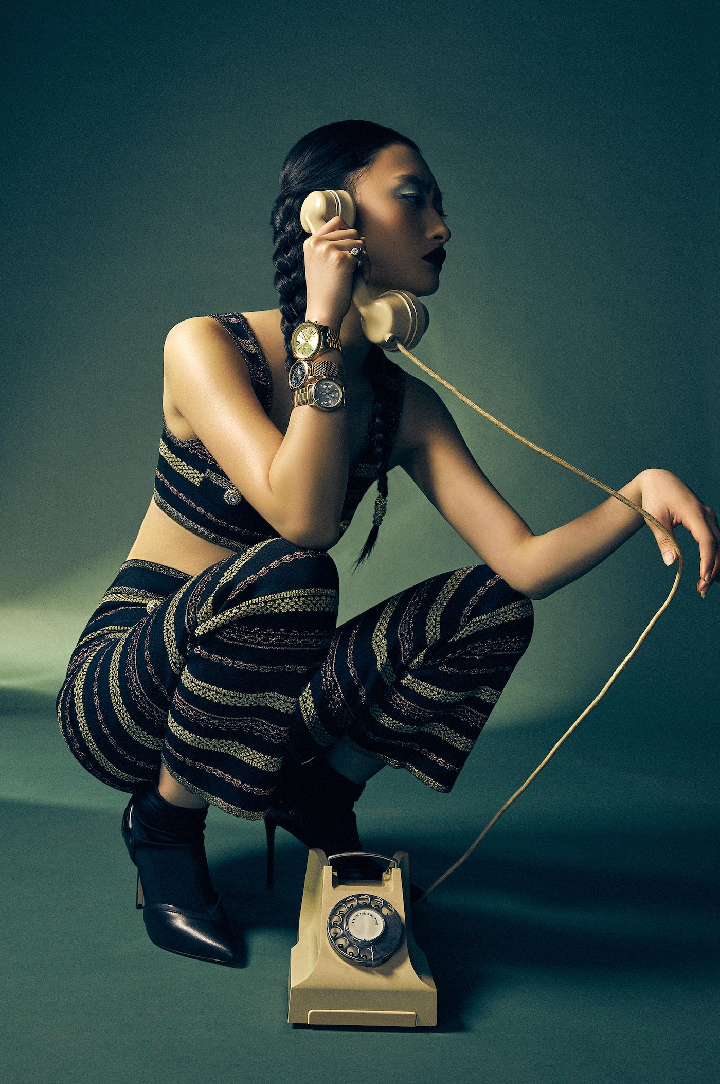 An Asian female fashion model crouches in a studio while holding a beige rotary phone. She wears a matching patterned top and pants.