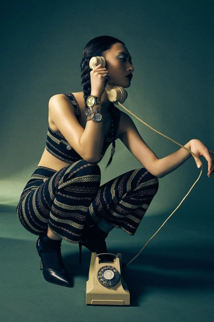 An Asian female fashion model crouches in a studio while holding a beige rotary phone. She wears a matching patterned top and pants.