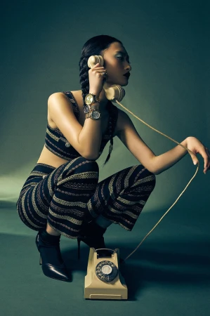 An Asian female fashion model crouches in a studio while holding a beige rotary phone. She wears a matching patterned top and pants.