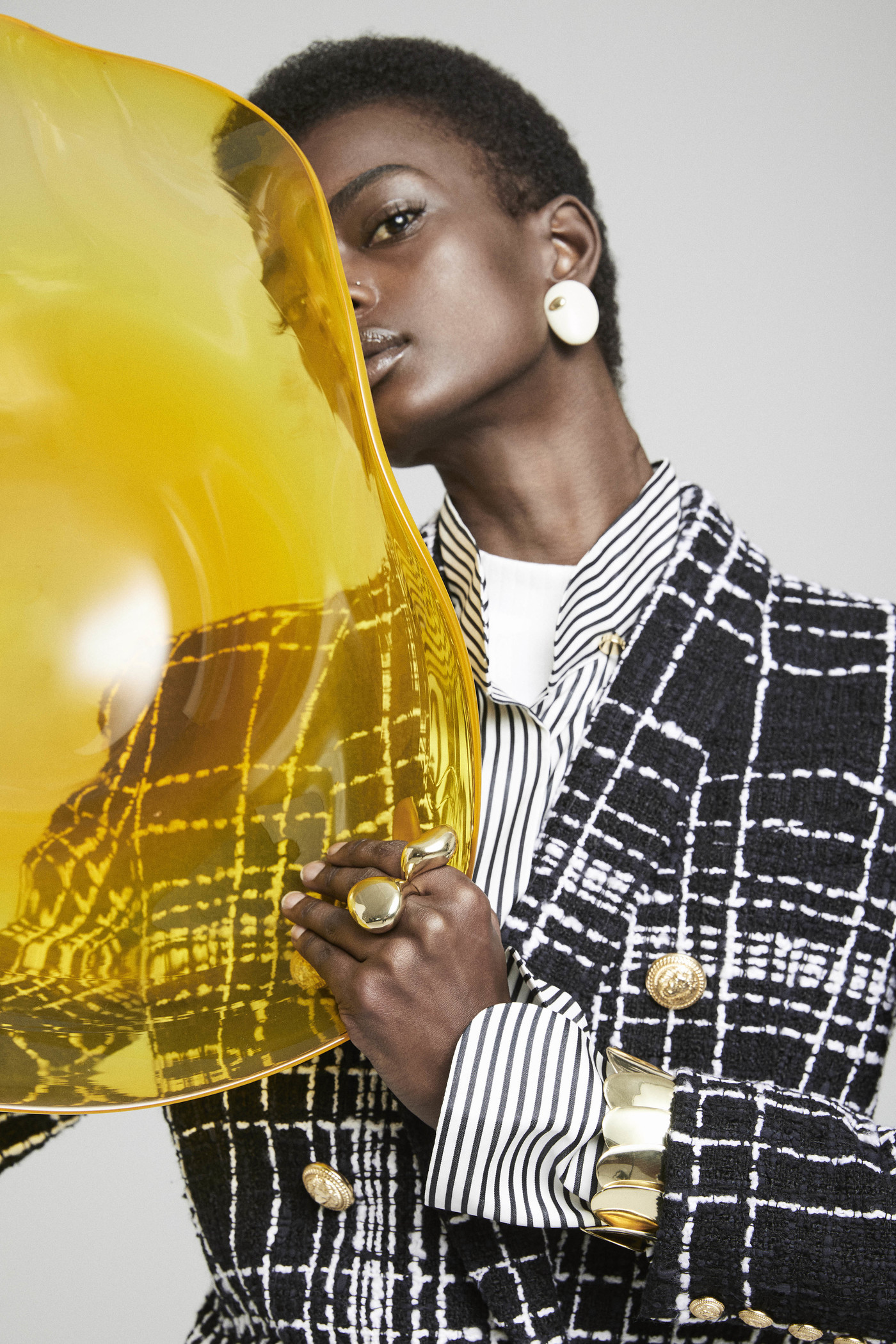 Black female model poses with a gold translucent shield, wearing a striped shirt, black and white jacket, chunky ring, and disc earrings.