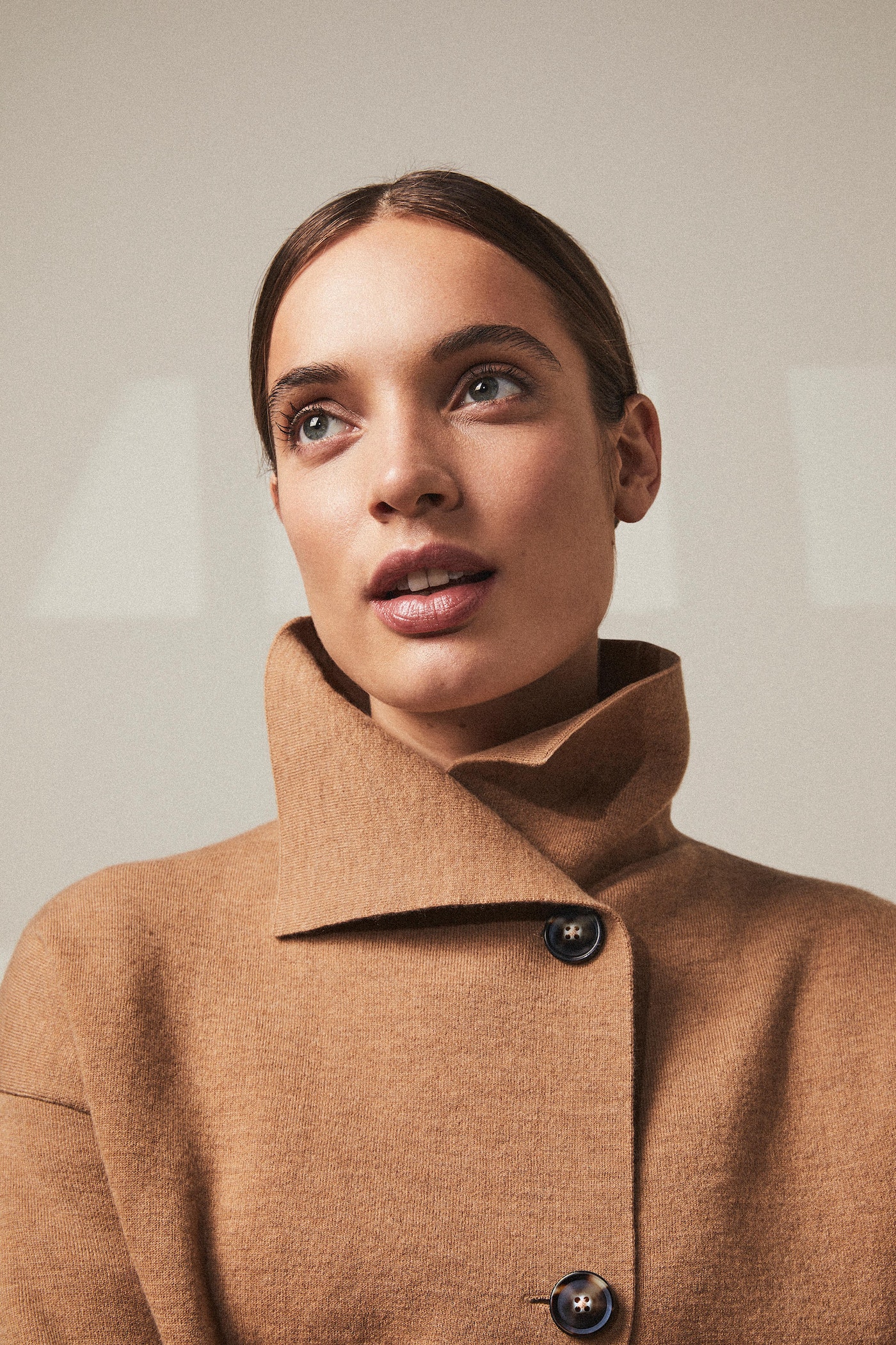 Portrait of a stylish woman in a tan coat looking up against a neutral background. Fashion and beauty photography.
