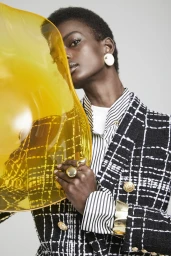 Black woman wearing black and white tweed jacket and striped shirt, holding large yellow curved glass prop