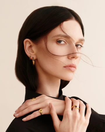 Close-up portrait of a woman with dark hair wearing gold rings and earrings against a plain light background