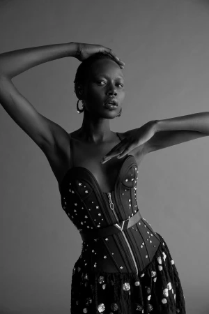 Black and white studio portrait of a Black model in a heavily embellished strapless corset top with both hands raised.