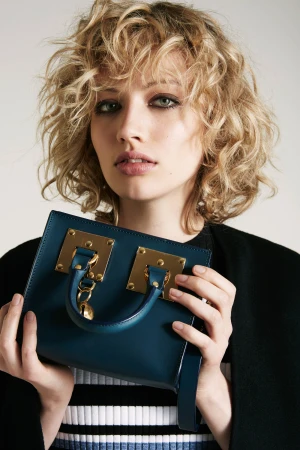 Woman with curly blonde hair holding a dark teal handbag with gold hardware against a plain white background.