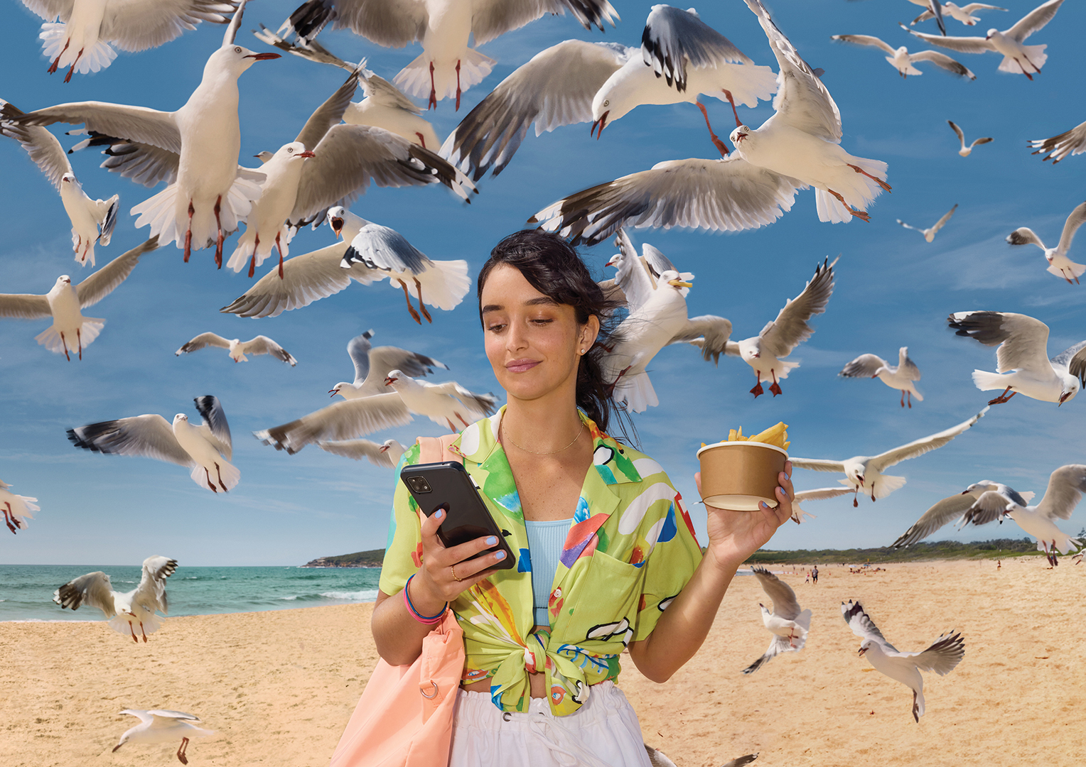 Woman in vibrant shirt checks her phone surrounded by seagulls on a sandy beach with a paper cup of fries in her hand.