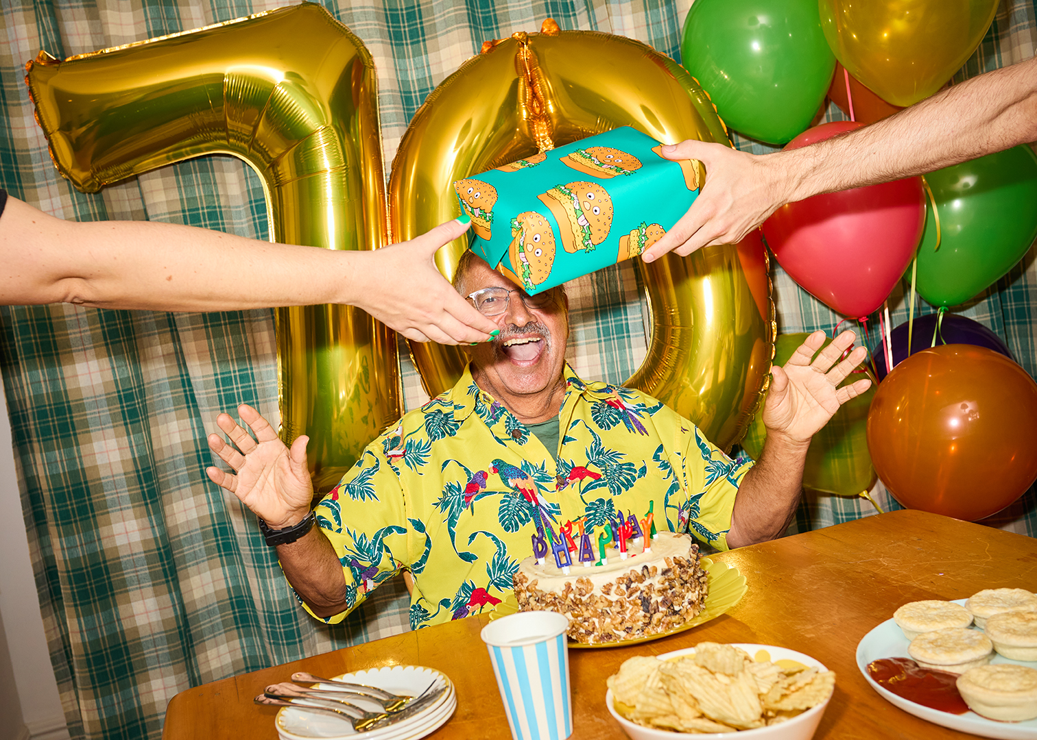 Mature man celebrating birthday with burger-themed gift, cake, and balloons in an indoor party setting with two unidentifiable people.