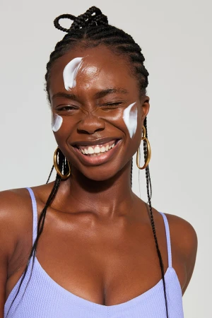 Black woman with skin cream on her face smiling while wearing a light purple top and gold hoop earrings