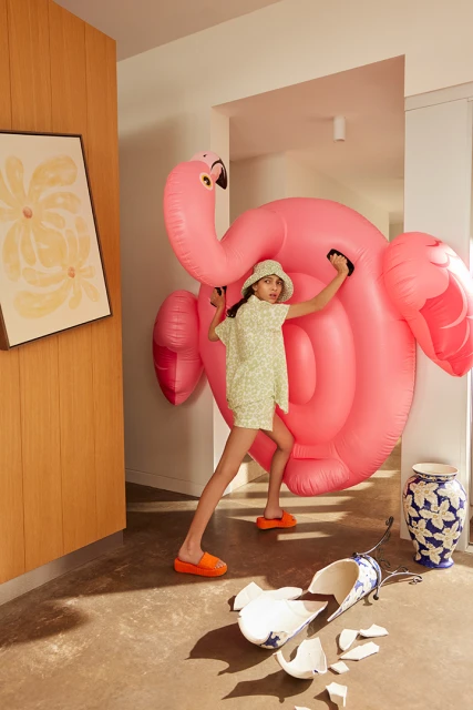 Child poses with a large inflatable flamingo in a hallway with a broken vase, wearing a floral shirt, shorts and matching bucket hat.
