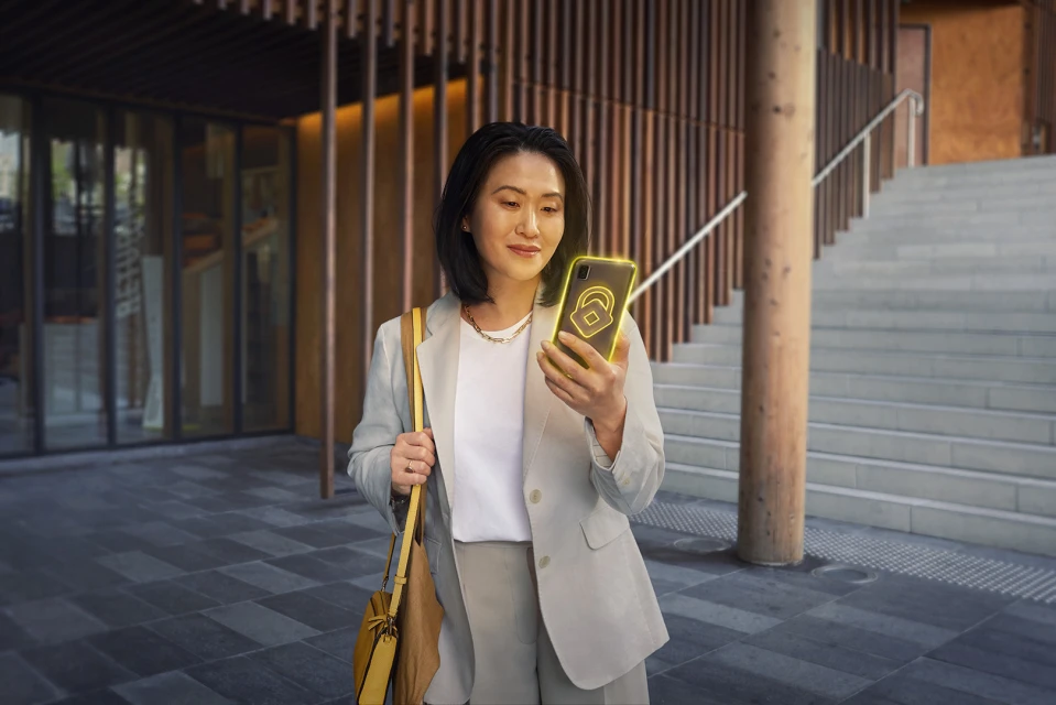 An Asian woman in a blazer is standing outdoors, holding a smartphone with a lock icon. She has a purse over her shoulder.