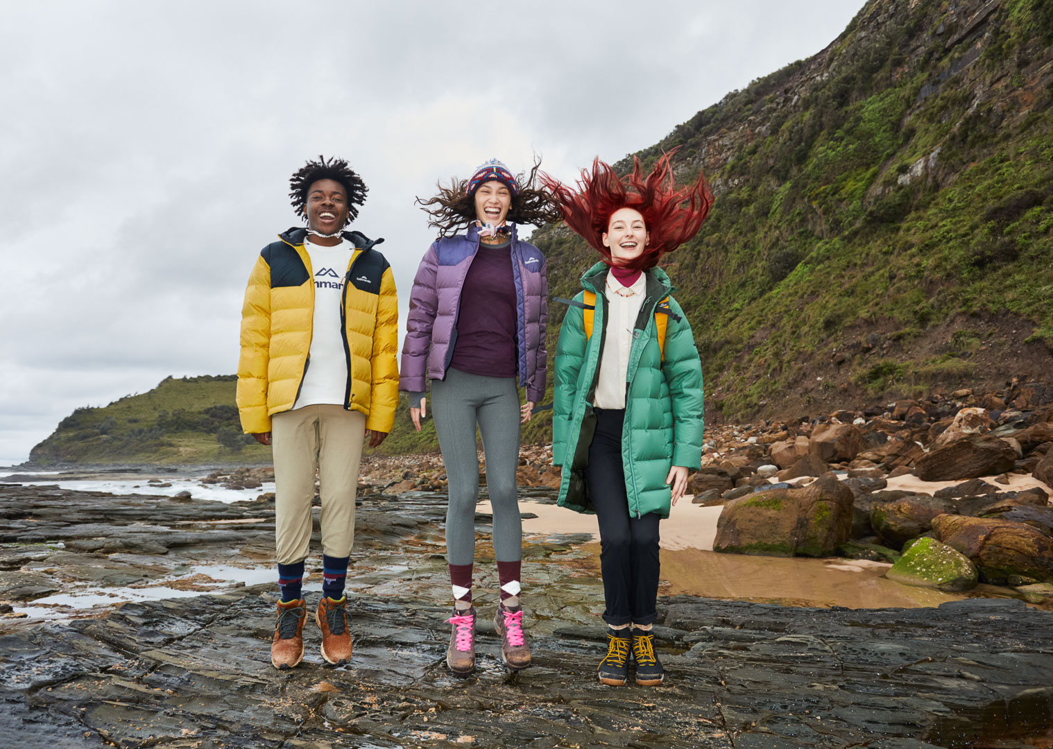 Three young adults jump on rocky terrain near the ocean, wearing puffy jackets; a cloudy day sets a sporty, adventurous mood.