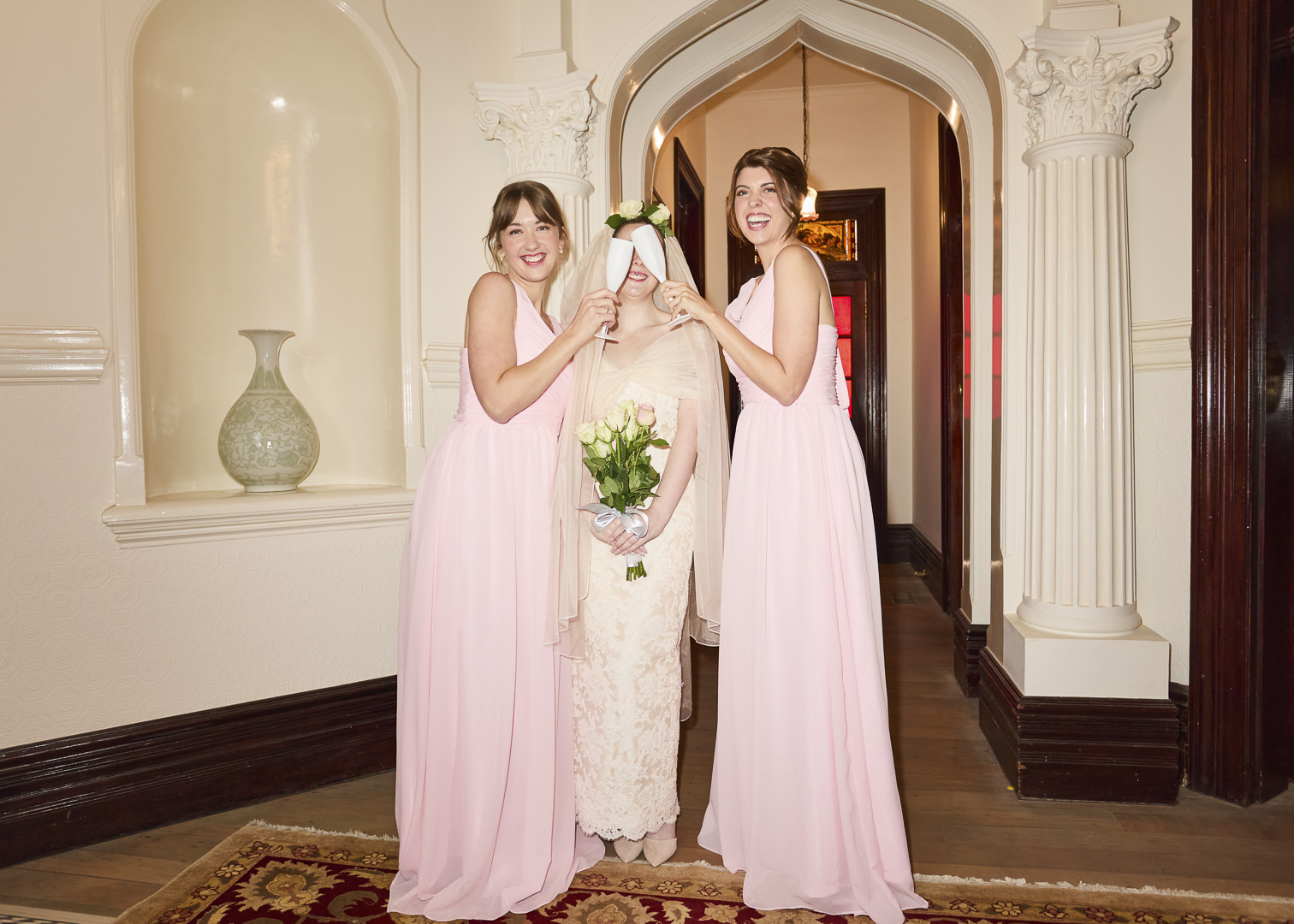 Bride with bridesmaids in pink dresses holding flowers in indoor venue with arched doorway and columns, celebrating wedding day.