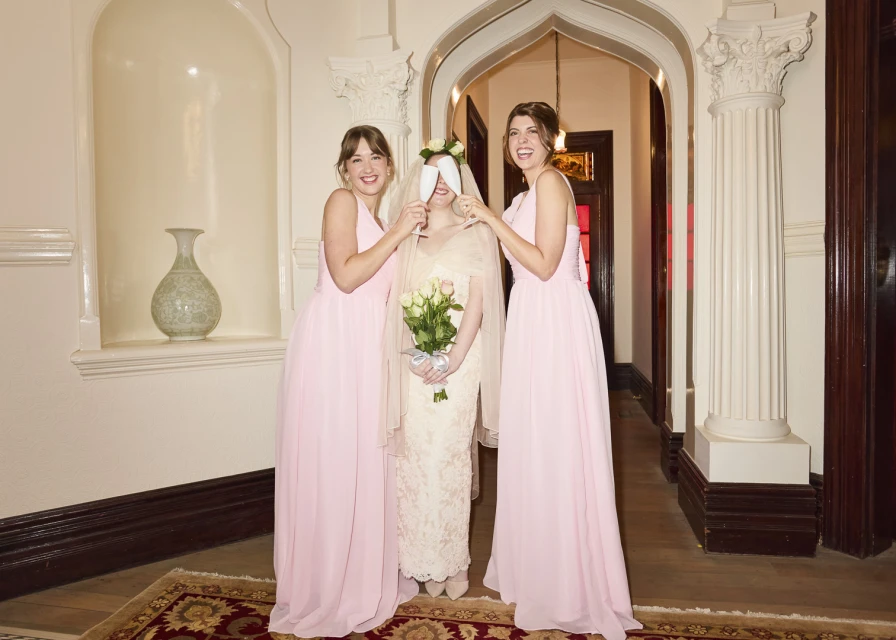 Bride with bridesmaids in pink dresses holding flowers in indoor venue with arched doorway and columns, celebrating wedding day.